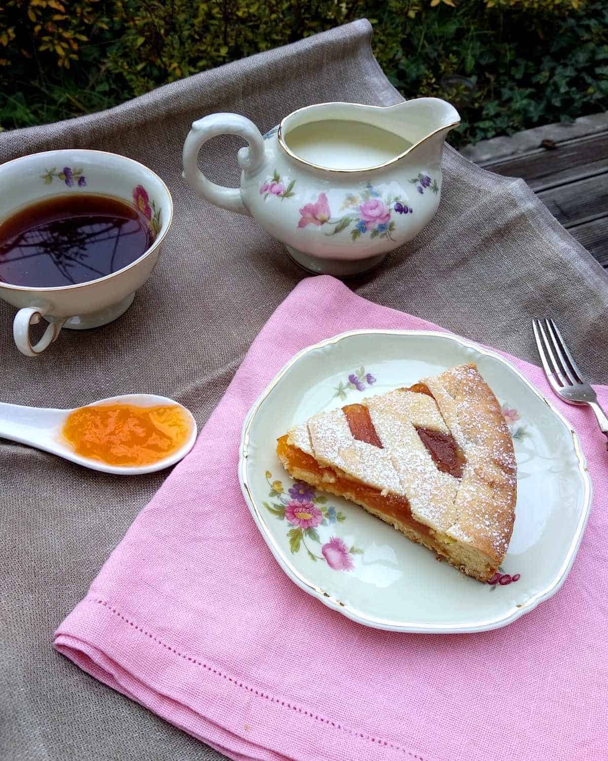 Una fetta di Crostata di Albicocche su un piatto bianco e lino rosa che mostra la marmellata di albicocche cotta all'interno della torta. Ci sono una tazza di caffè e un po' di latte da parte