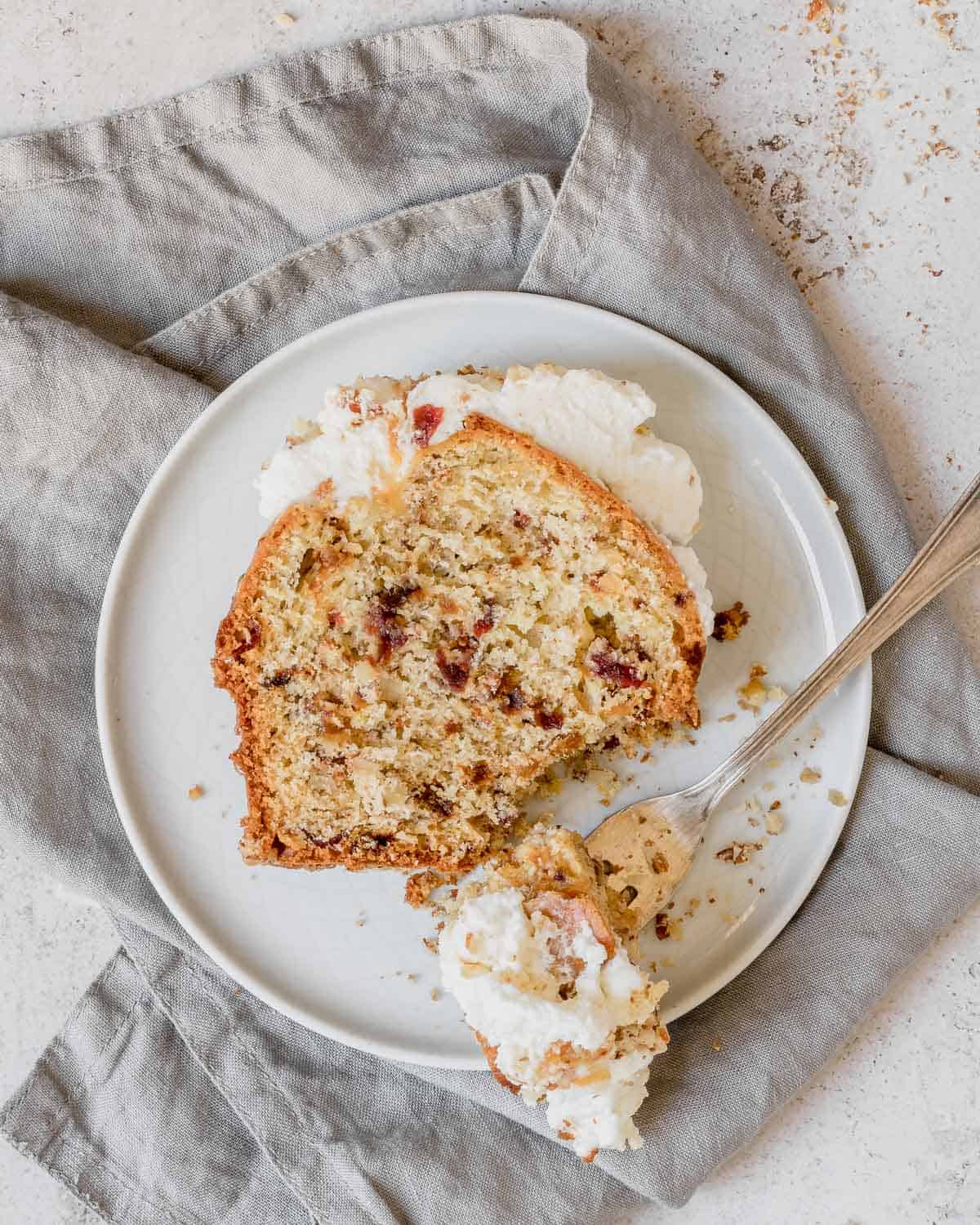 Una fetta di plumcake alla frutta disidratata con noci e mandorle-su-un-piatto-bianco-che-mostra-i-frutti-all'interno-del-plumcake-è-guarnito-con-panna-montata-fresca-e-caramello-C'è-la-forchetta-con-un-pezzo-di-torta-sul-piatto-