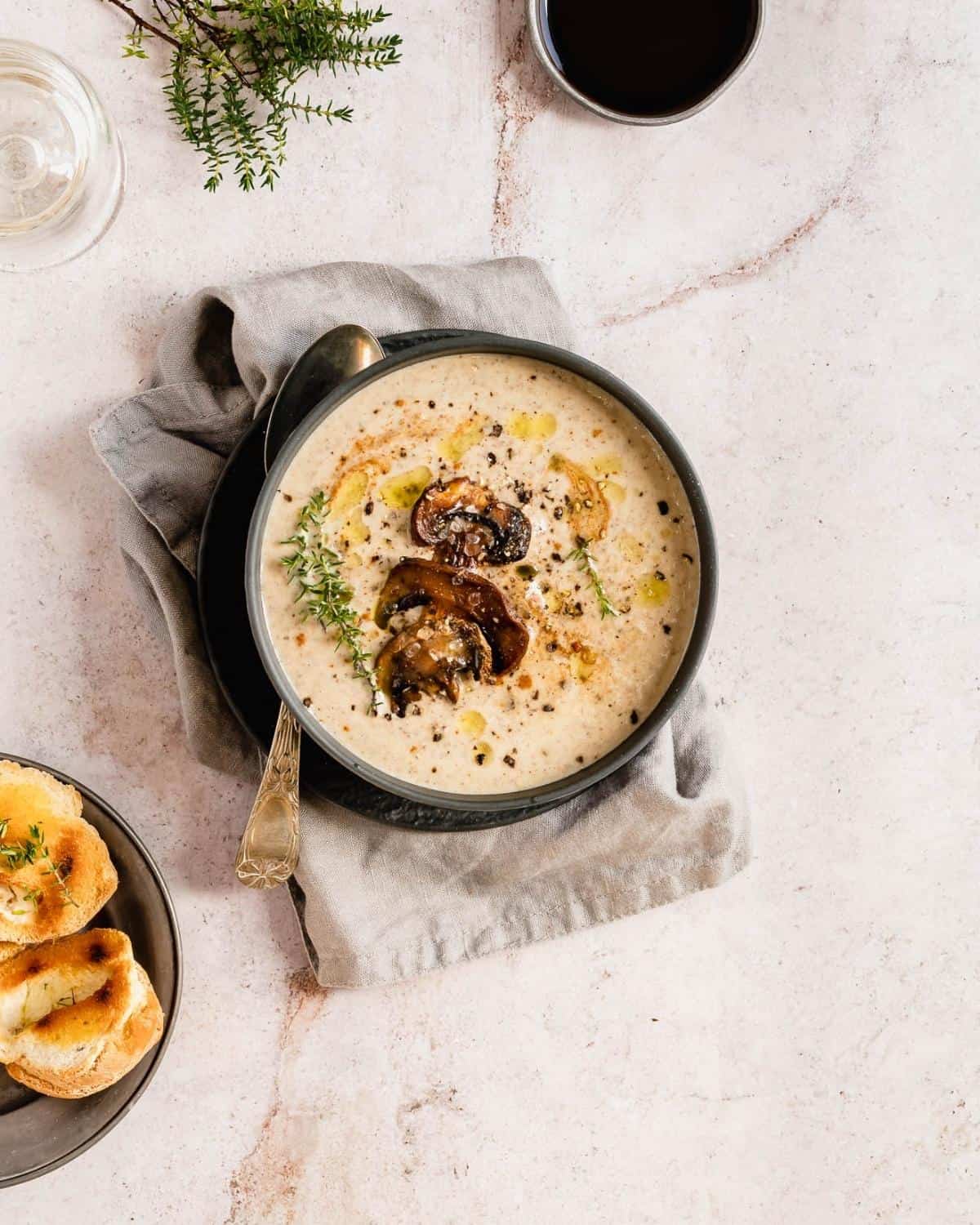 Zuppa di funghi in una ciotola scura su un tavolo di lino e marmo beige. Il piatto è guarnito con funghi e pepe nero. A parte un cucchiaio e delle fette di pane abbrustolite