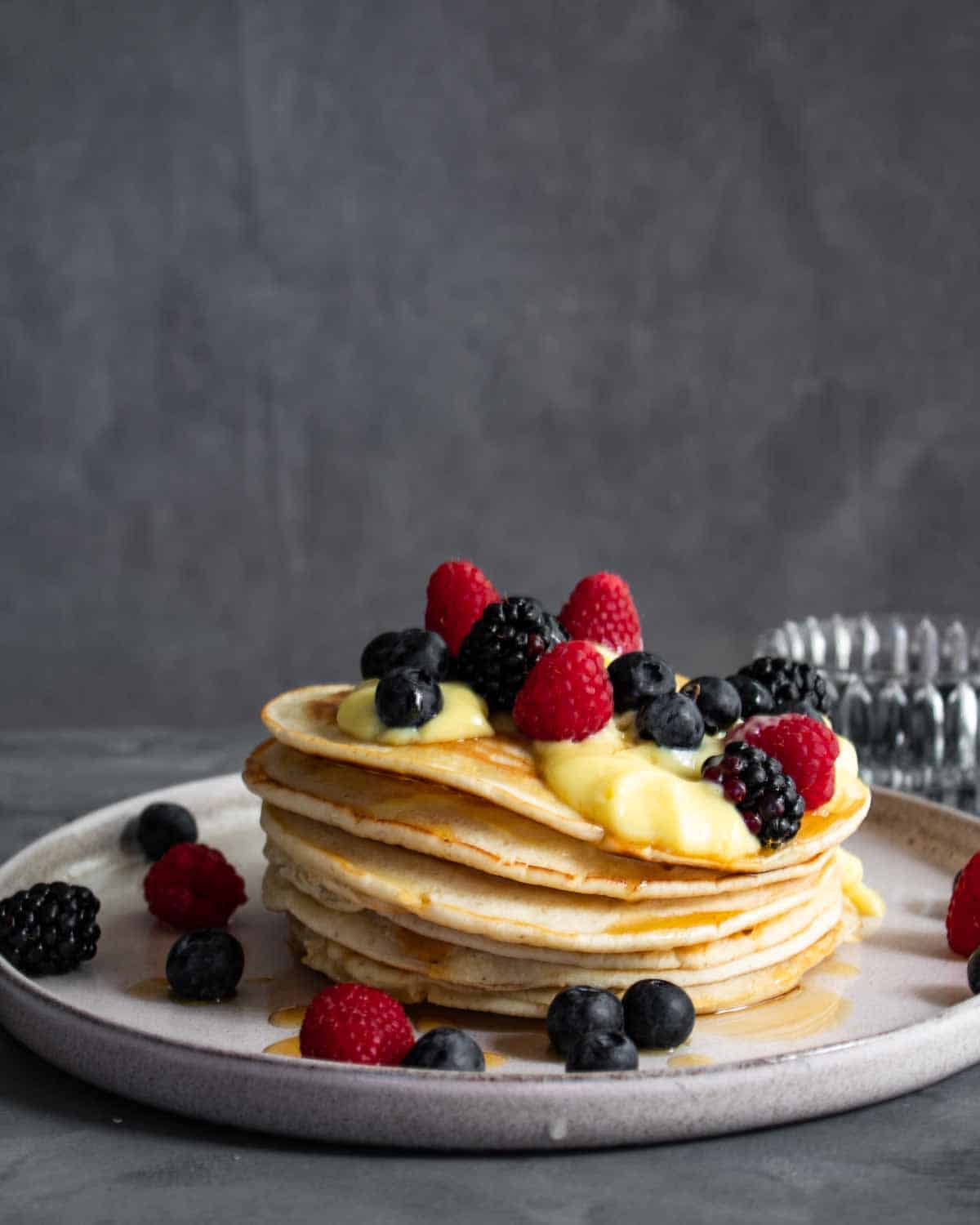 Una foto dritta di una pila di pancakes su un piatto bianco e un tavolo grigio. Sono farciti con crema pasticcera e frutti di bosco