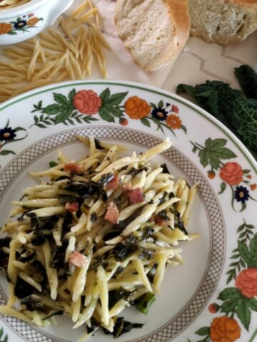 A view from above of Black Kale Pasta in a white plate with flowers. The dish is garnished with Pancetta and Parmesan cheese