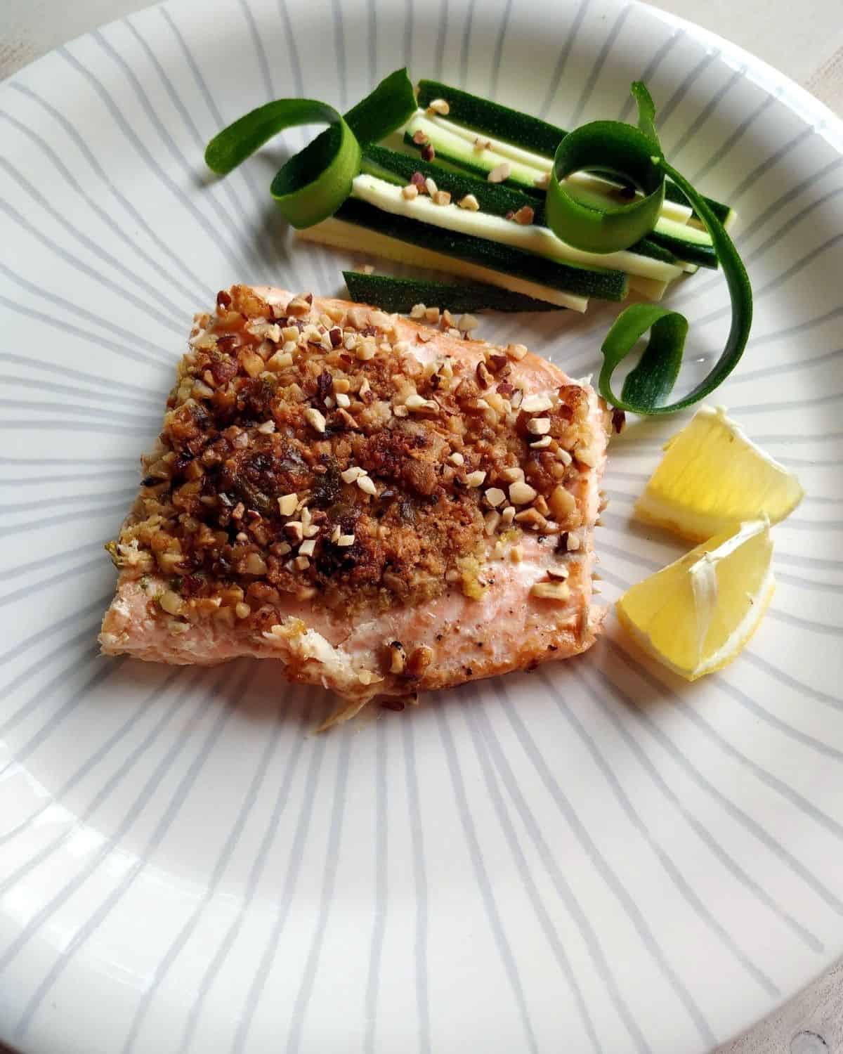 A view from above of Almonds Crusted Salmoni in a white dish showing the almond crust on top. It is served with sliced zucchini and lemon.