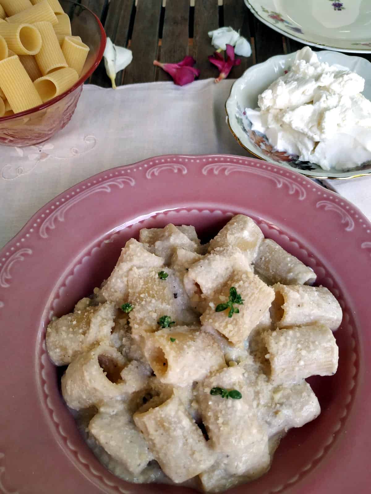 A Onion and Ricotta Pasta on a pink plate and white linen shown from above. A little plate with ricotta on the background
