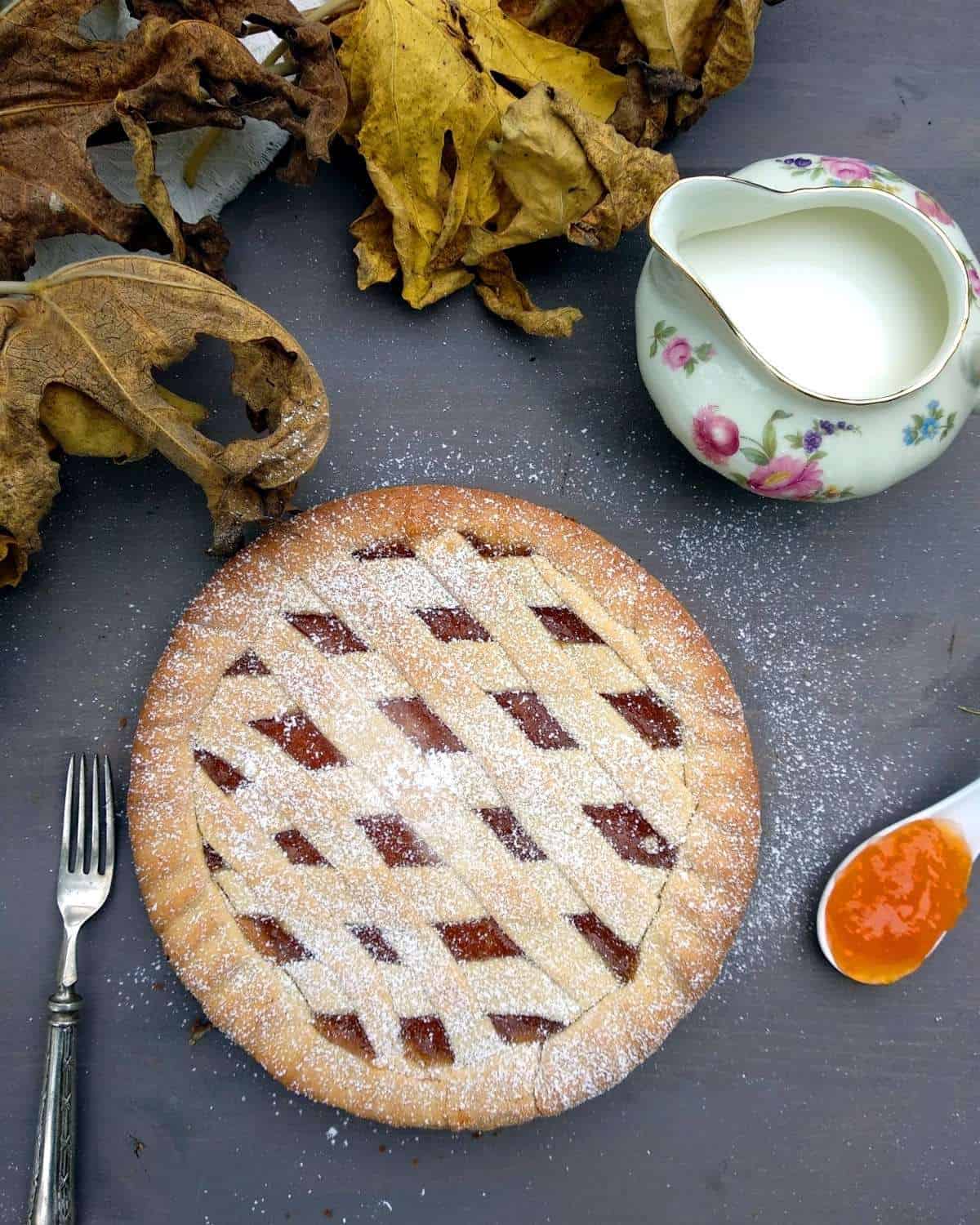 A close up shot from above of the Apricot Crostata showing the lattice with apricot jam. The tar is on a blue surface with a spoon full of jam on the right side and a fork opposite side