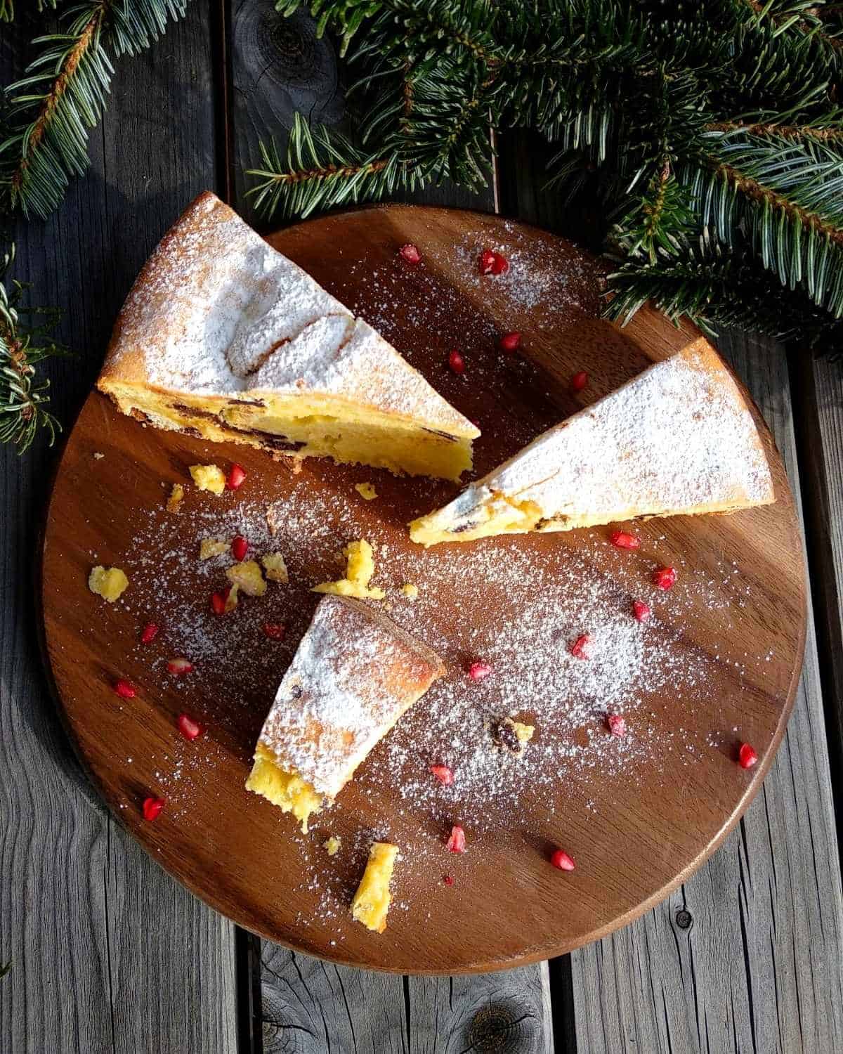 A close up from above Ricotta and Chocolate Cake on a wooden board. It is sliced showing the chocolate inside the cake. Some Pine branches in the background