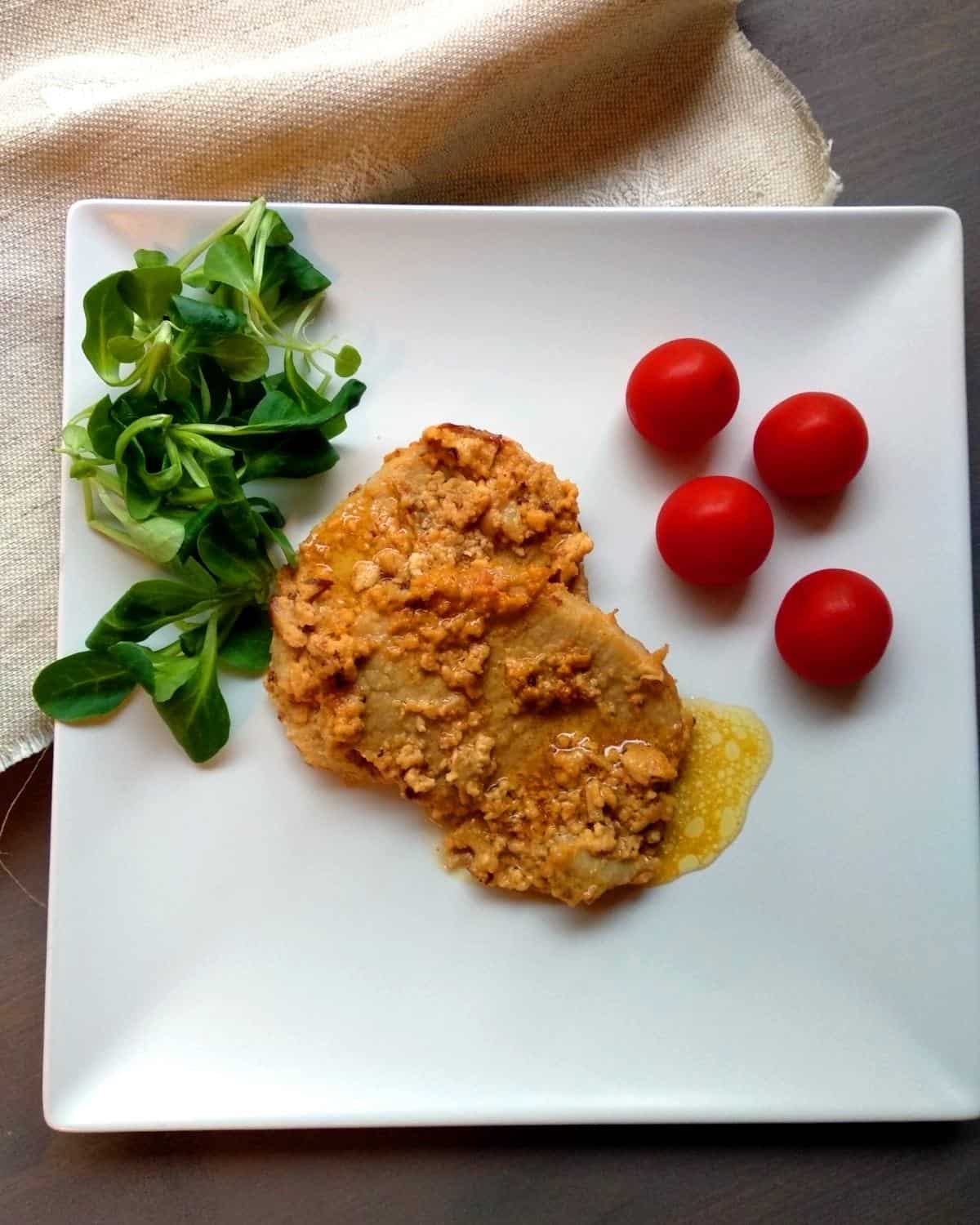 A slice of Pork Braised in Milk on-a-white-dish and light brown linen. It is showing the rich and creamy sauce. There is fresh salad and cherry tomatoes in the plate-