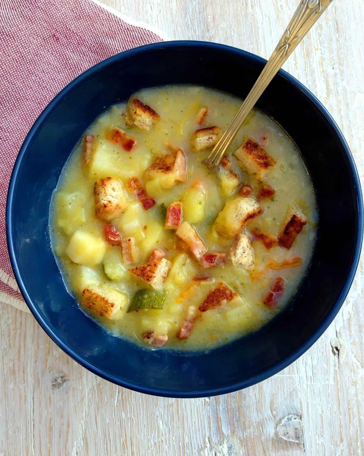 A blue bowl of Potatoes and Bacon Soup on a red linen and white table. There is a spoon inside the soup