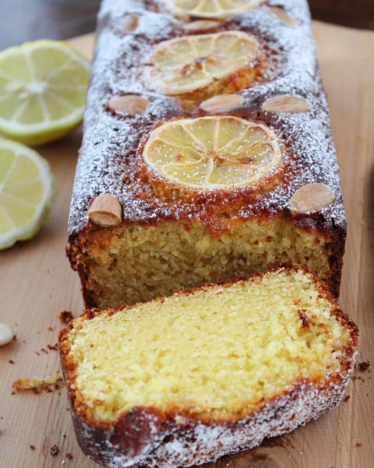 An almond Loaf on a wooden board. The cake is sliced and there are lemon slices and almonds on top