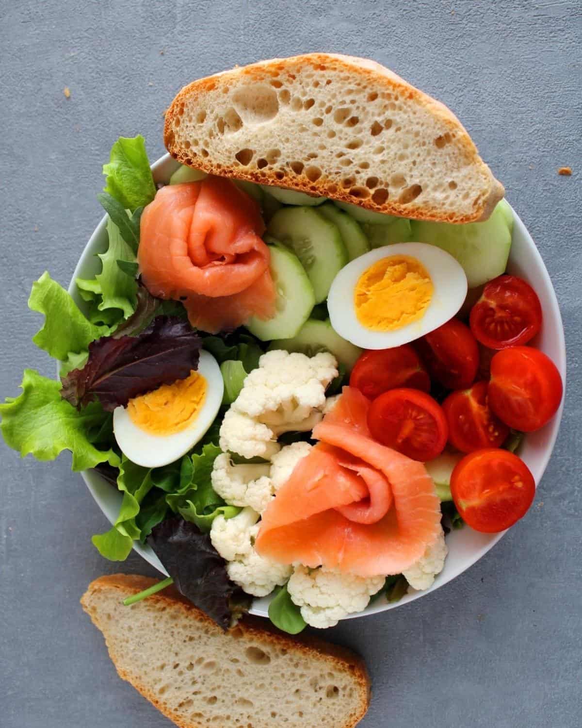Top view of a bowl of a salmon cucumber cauliflower salad and an egg cut in half. Two slices of bread
