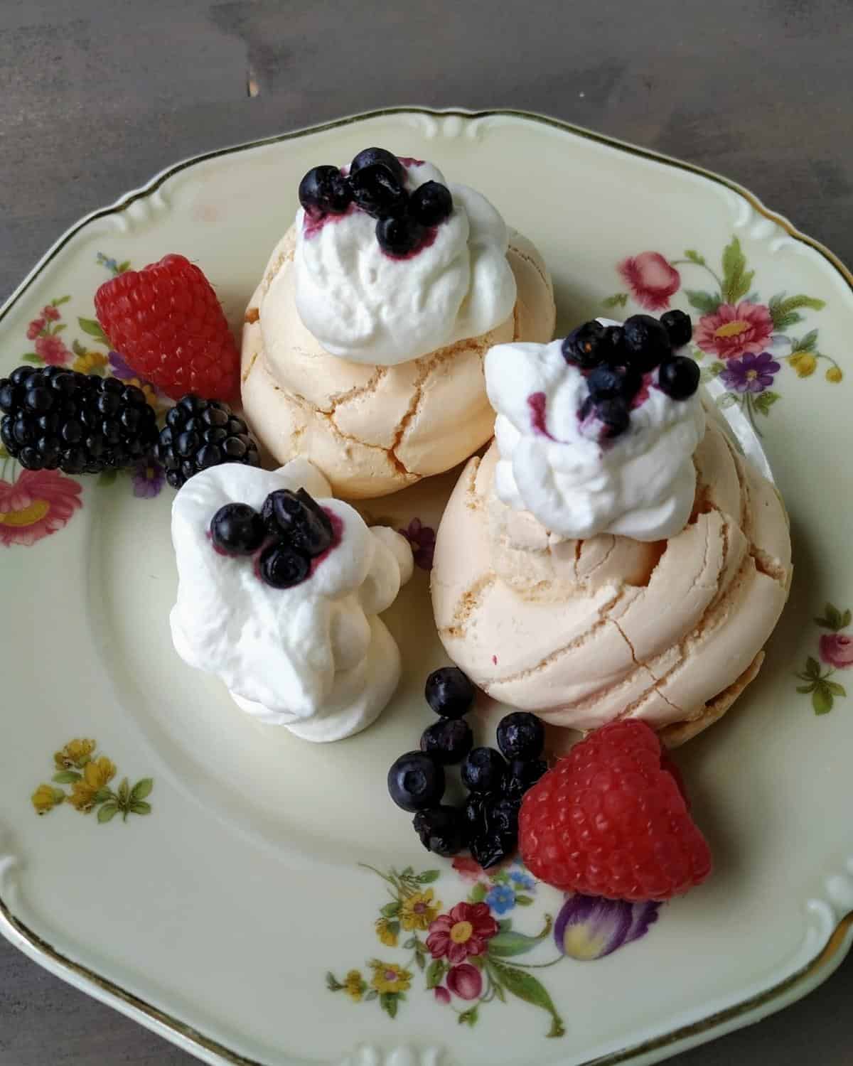A close up from above photo of three meringue on a white floured plate. They show whipped cream and wild berries on top