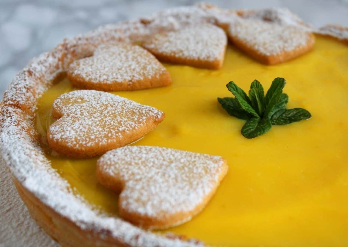 A close up of Lemon Curd Tart. It shown the lemon curd and three hearts of dough. The crostata is topped with icing sugar and some mint leaves