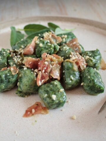 Gnocchi with Spinach in a light dish with a fork on a red linen. Gnocchi are garnished with speck and grated parmesan