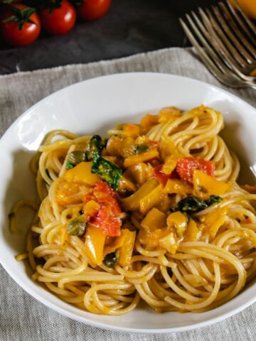 A 45 degree view of Pasta with Peppers in a white dish on a beige linen. It shown peppers sauce with cherry tomatoes and fresh basil leaves