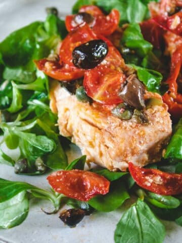 A salmon fillet on a white plate. It is garnished with black olives, cutted tomatoes and salad
