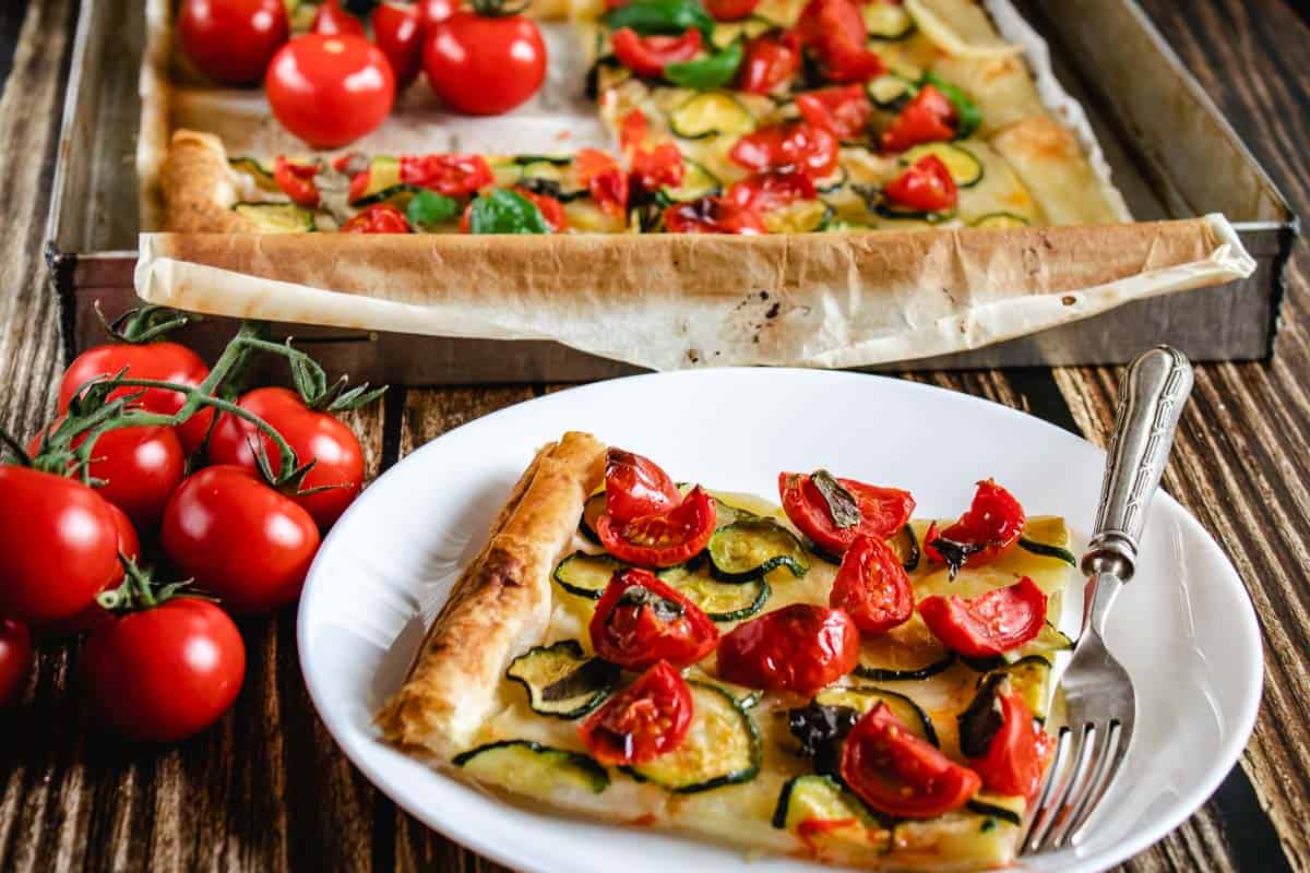 A close up a Zucchini Tart slice in a white plate with a fork on a wooden surface. The slice shown zucchini, potatoes and cherry tomatoes baked on top. In the background there is the tart