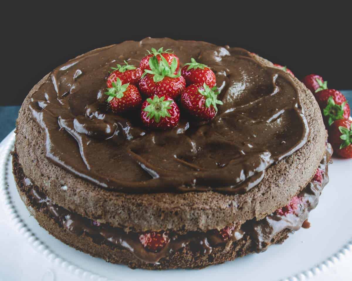 A close up of Chocolate and strawberries Cake on a white cake stand. It shown the chocolate and strawberries between the two layers. It is topped with a lot of chocolate and strawberries