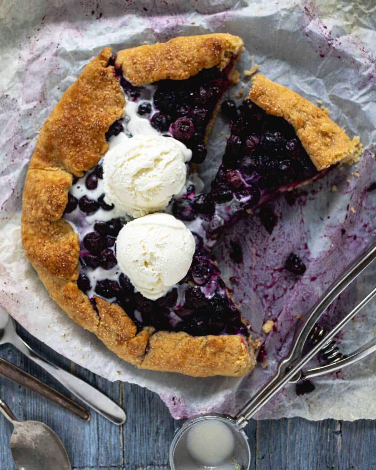 A Blueberries Galette on a blue rustic table with parchment paper shown from above. The galette has a lot of blueberries baked inside and two ice-cream balls on top