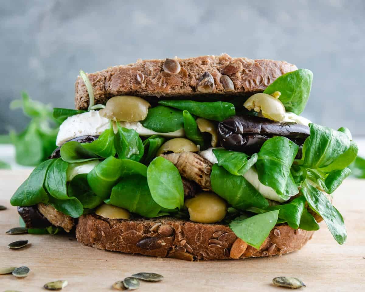 A Eggplants and Mozzarella Sandwich close up in a light wood board. The sandwich shown layers of eggplants, salad mozzarella and green olives