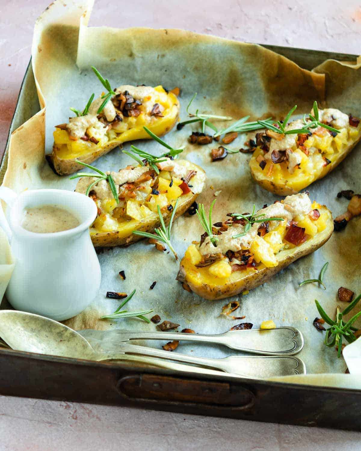 4 half cut potato filled with mushrooms cheese and bacon on a baking tray with parchment paper. A white bowl with cheese cream and forks and spoon on the left