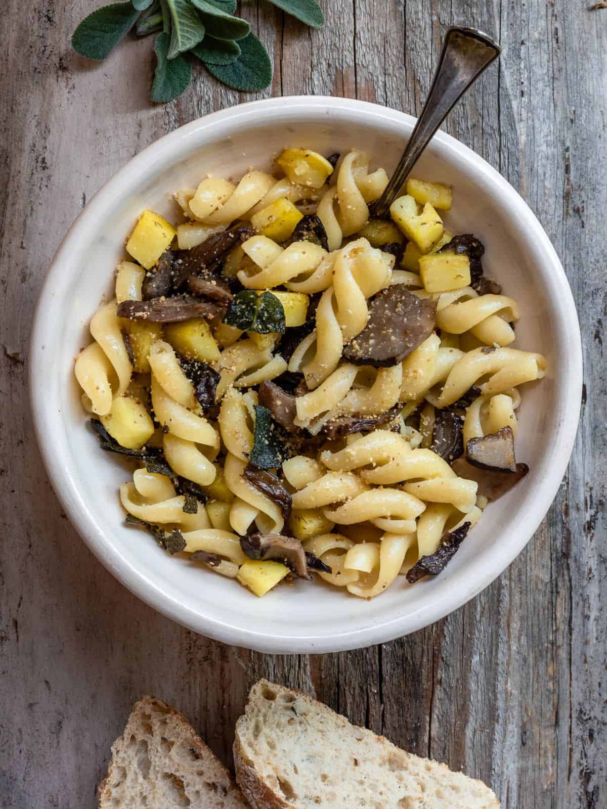 A view of Mushrooms Pasta with Potatoes and Sage in a light bowl on a rustic wooden table. The dish shown mushrooms, potatoes and browned sage on top. On the side some fresh sage and 2 slices of bread