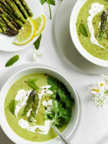 Two white bowls of Asparagus Soup on a white table. There is a spoon inside the soup. On the side a little plate with few asparagus