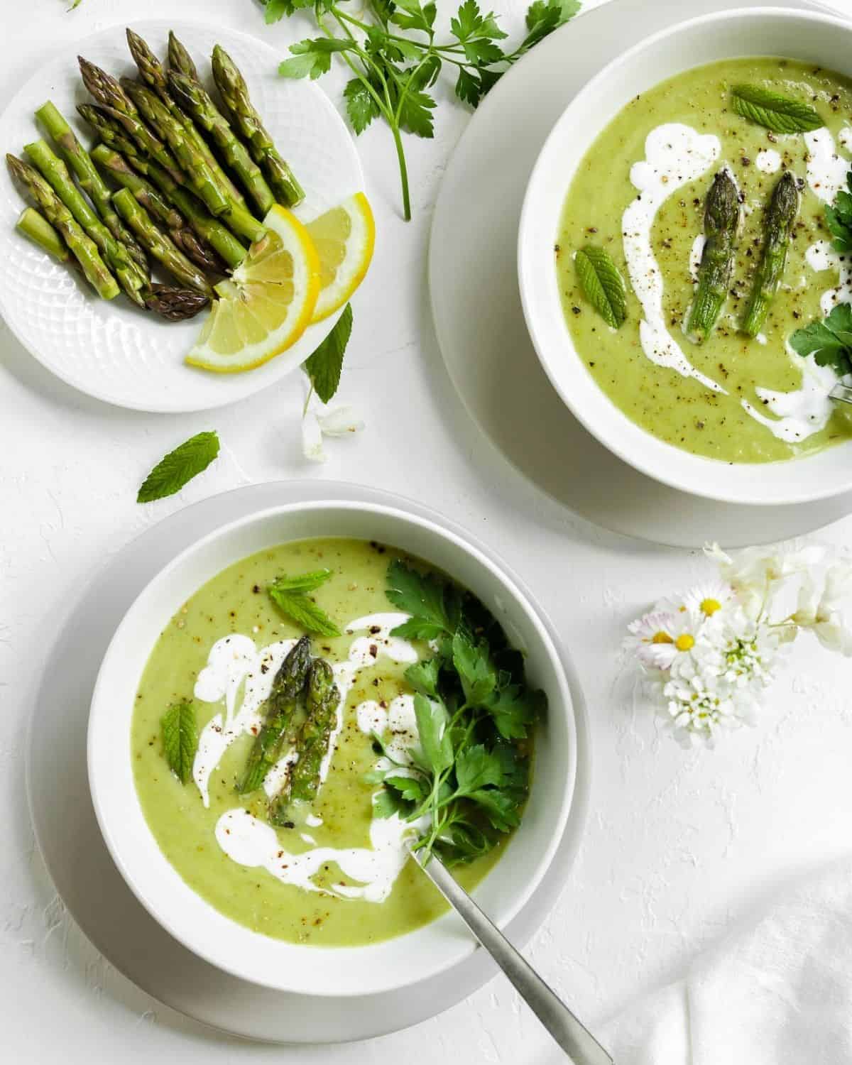 Two white bowls of Asparagus Soup on a white table. There is a spoon inside the soup. On the side a little plate with few asparagus