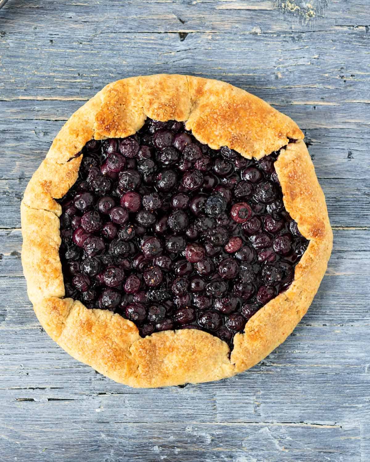 A Blueberries Galette on a blue rustic table shown from above. The galette has a lot of blueberries baked inside