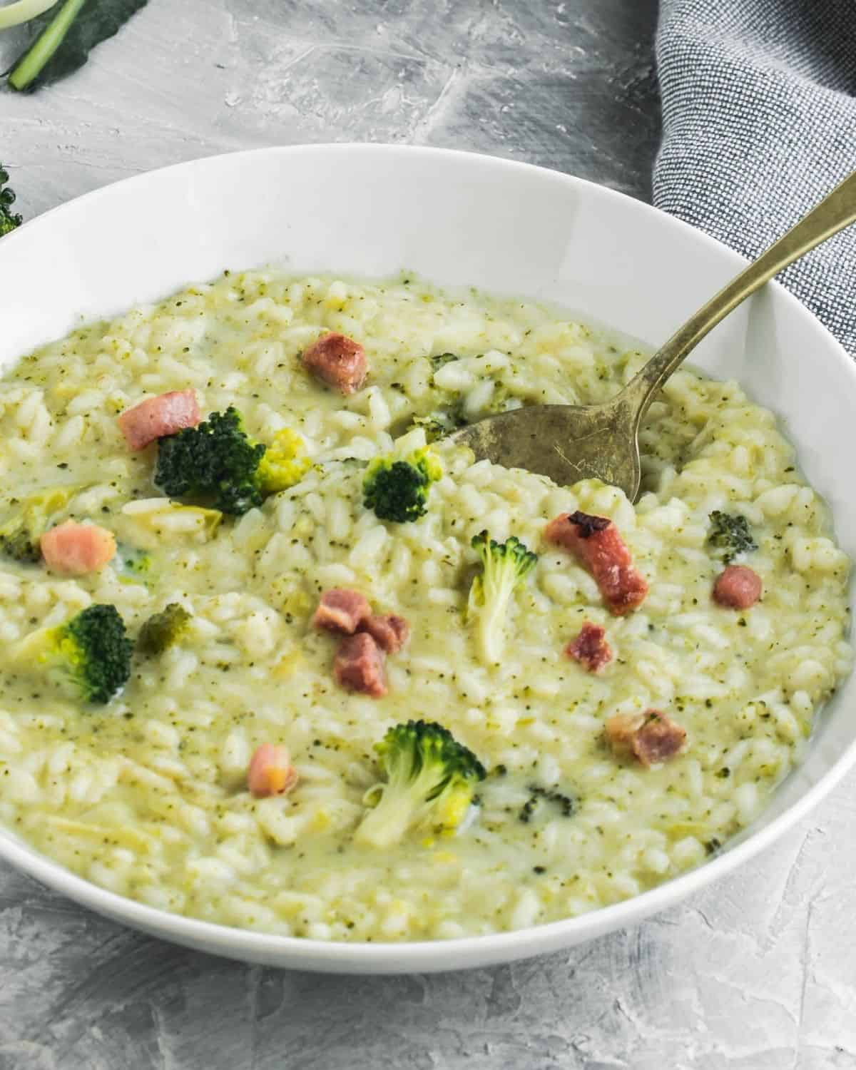 A close up of Broccoli Risotto with Gorgonzola in a white plate with a fork. It is topped with broccoli florets  and pancetta