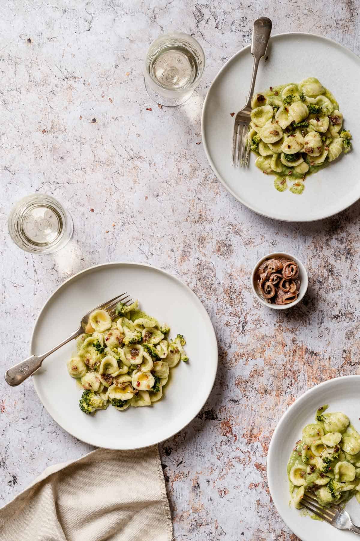 A close up from above of 3 Broccoli Orecchiette Pasta on white plates and marble table. The pasta shown a few anchovies on top. There are 2 glasses of white wine and a little bow of anchovies.