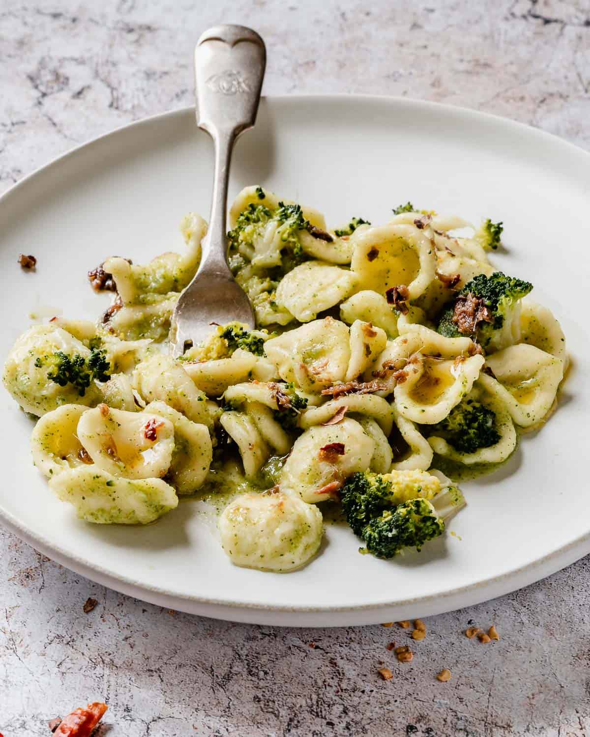 A close up of Broccoli Orecchiette Pasta on a white plate and marble table. The pasta shown a few anchovies on top. There is a fork in the plate