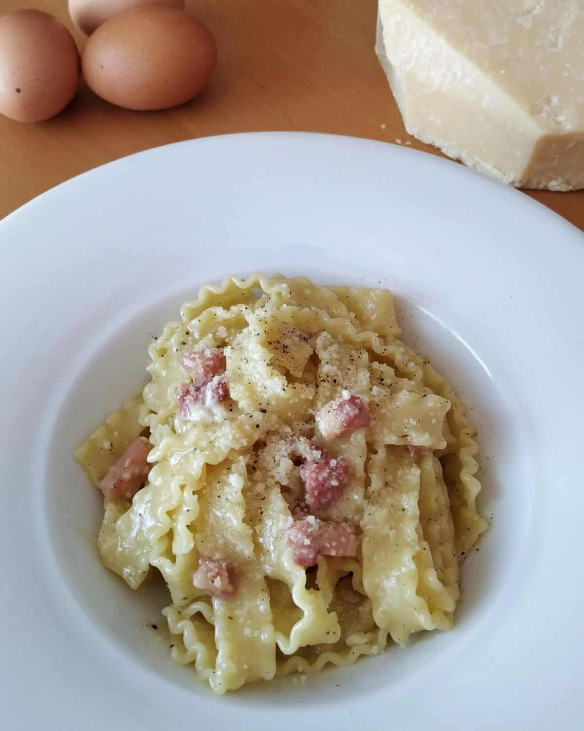 A close up of Carbonara Pasta on a white plate. Eggs and cheese in the background
