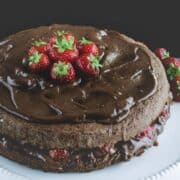A close up of Chocolate and strawberries Cake on a white cake stand. It shown the chocolate and strawberries between the two layers. It is topped with a lot of chocolate and strawberries