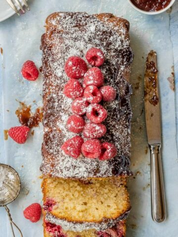 A-Coconut-and-Raspberry-Loaf-on-the-light-blue-table-with-parchment-paper-the-loaf-has-fresh-raspberries-and-icing-sugar-on-top- it-is-sliced-showing-the -raspberries-inside-there-are-a-bowl-of -jam-and-a knife-aside