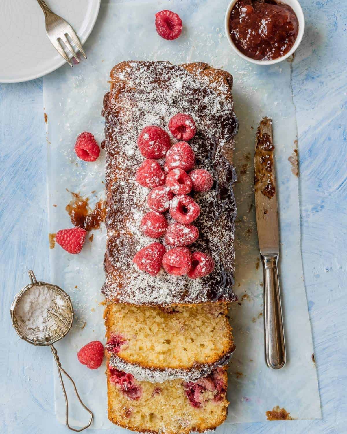 A-Coconut-and-Raspberry-Loaf-on-the-light-blue-table-with-parchment-paper-the-loaf-has-fresh-raspberries-and-icing-sugar-on-top- it-is-sliced-showing-the -raspberries-inside-there-are-a-bowl-of -jam-and-a knife-aside