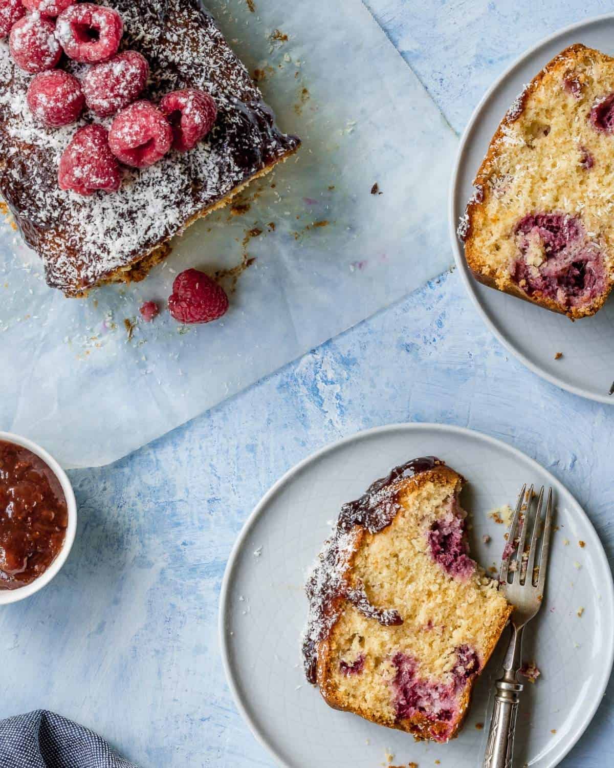 A-Conut-and-Raspberry-Loaf- showing-a-lot-of-Raspberry-on top-2 slices of-Loaf-with-lots-of-Raspberry-baked-inside-the-slices-are-on-light-blue-plates-with-fork-and-jam-raspberrie-aside
