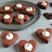A few Chocolate Hearts Cookies. Some are on a grey plate other on a rustic grey table. All cookies are topped with whipped cream