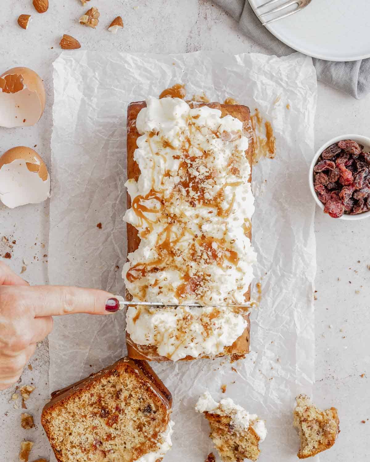 someone-is-slicing-a-Dried Fruits Loaf with Walnuts and Almonds-on-light-white-table-with-parchment-paper-the-loaf-has-fresh-whipped-cream-and-caramel-on-top- it-is-sliced-showing-the-dired-fruits-inside