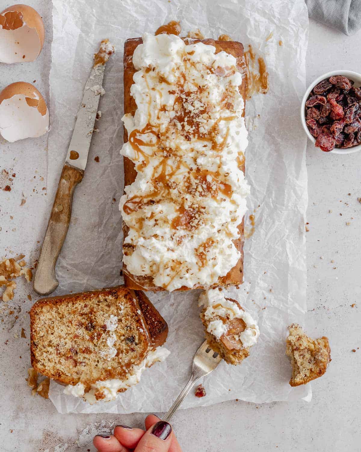 A-Dried Fruits Loaf with Walnuts and Almonds-on-the-light-white-table-with-parchment-paper-the-loaf-has-fresh-whipped-cream-and-caramel-on-top- it-is-sliced-showing-the-dired-fruits-inside-An-hand-keep-a-fork-with-a-piece-of-cake-there-are-a-bowl-of-dried-fruits-and-a knife-aside