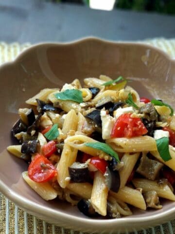 A close up of Eggplants Pasta on a white brown plate. The pasta shown few vegetables and a few basil leaves on top