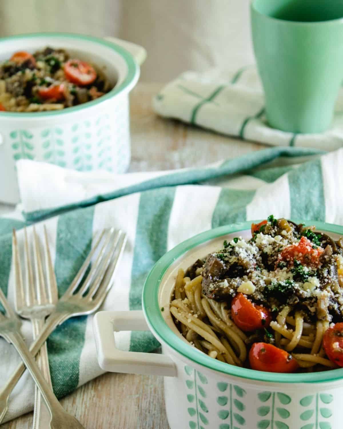 Two bowls of Eggplants Spaghetti with Anchovies on a white wooden table with a white and green linen. There are two forks and a green mug