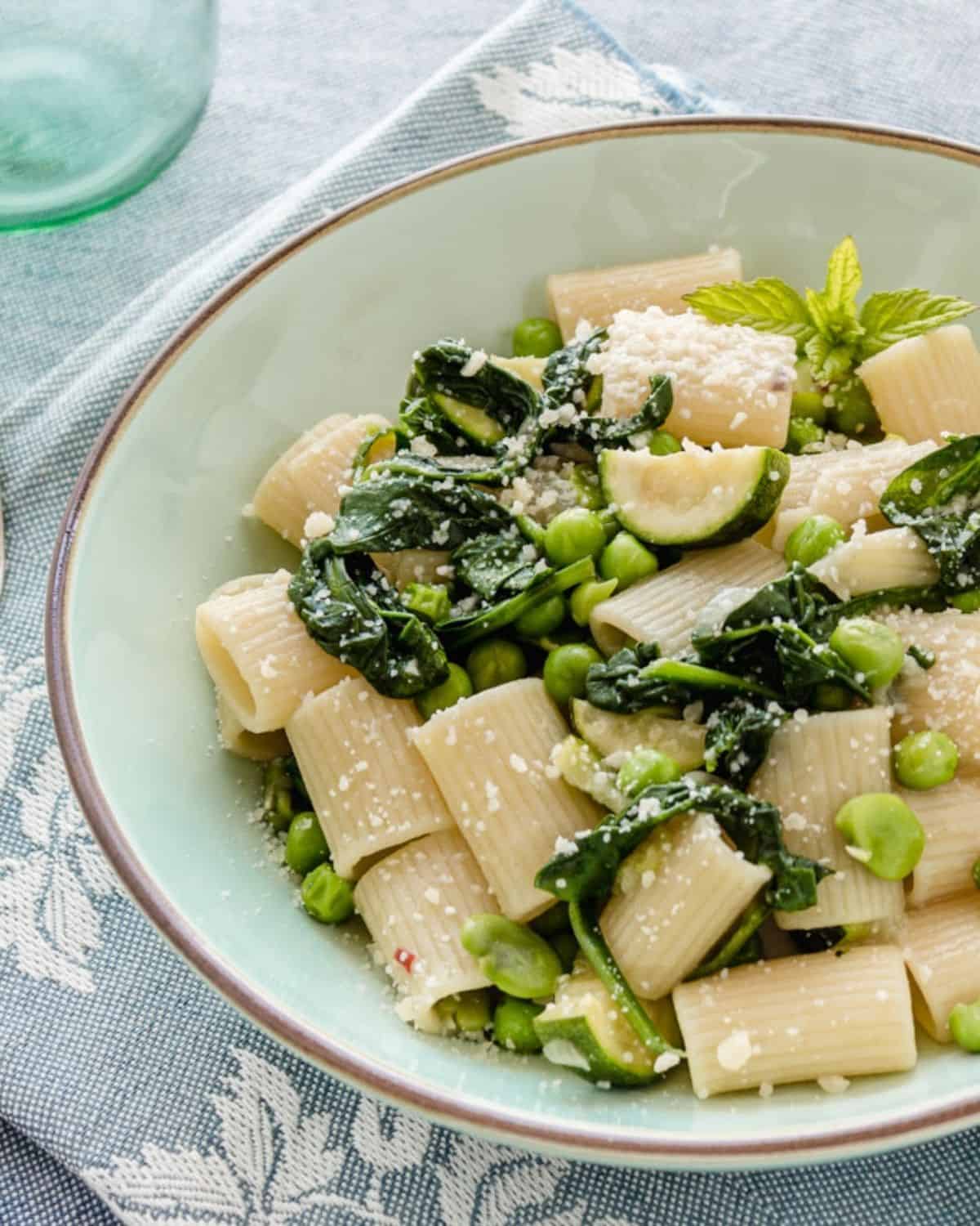 A close-up of Green Vegetable Pasta in a light green plate on blue linen. The dish shows spinach, peas and zucchini