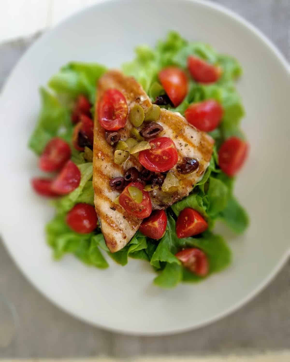 A Grilled-Swordfish  on a white plate. It is garnished with chopped tomatoes, olives and salad