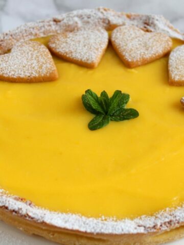A close up of Lemon Curd Tart. It shown the lemon curd and three hearts of dough. The crostata is topped with icing sugar and some mint leaves