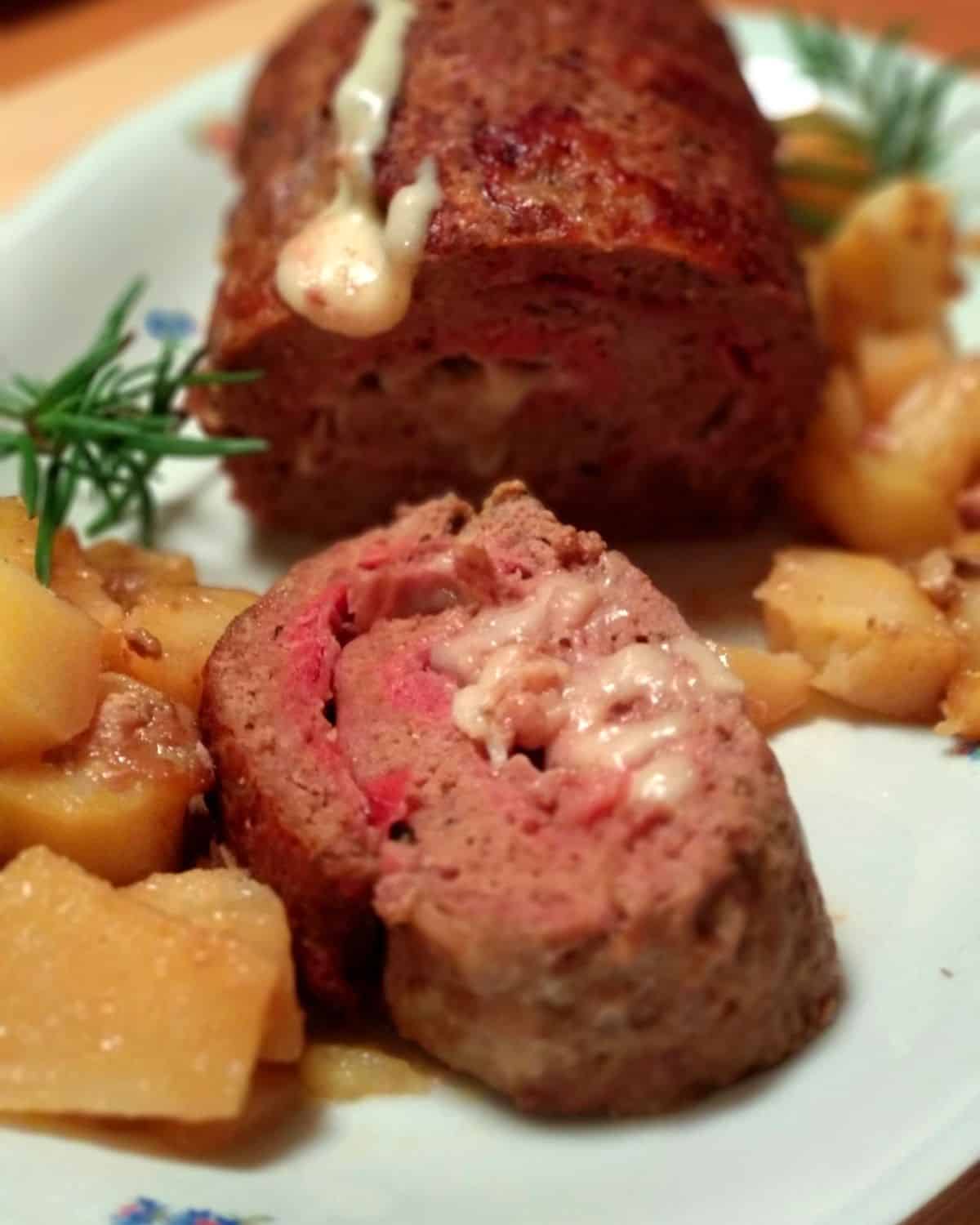 A Meat Loaf with Prosciutto on a white plate. It-is-sliced-showing-the -prosciutto and cheese layers inside. There are some roasted potato aside