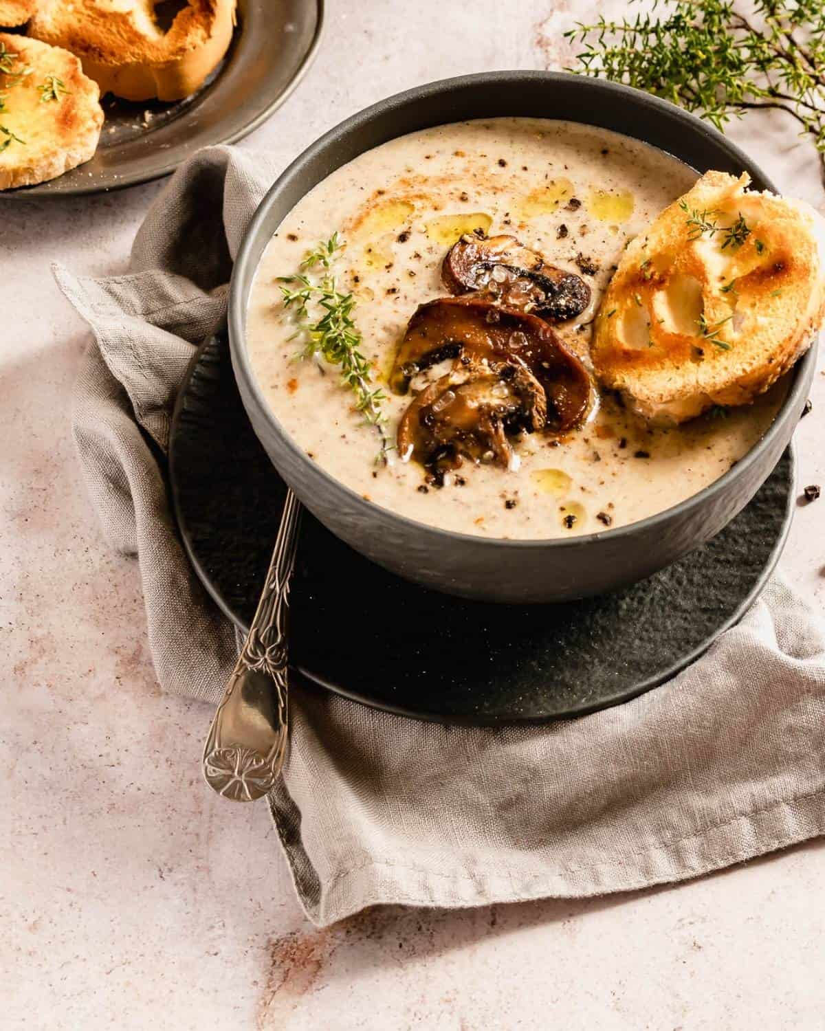 A Mushrooms soup in a dark bowl on a beige linen and marble table. The dish is garnished with Mushroom and black pepper. On the side a spoon and some roasted slices of bread