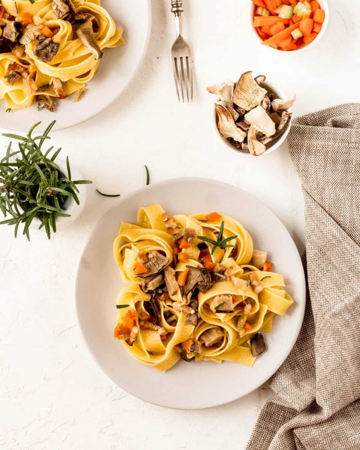 Two plates of Mushrooms and Bacon Tagliatelle on white plates. The pasta shown mushrooms and carrots on top. There are fresh rosemary in a glass and a bowl with mushrooms ns