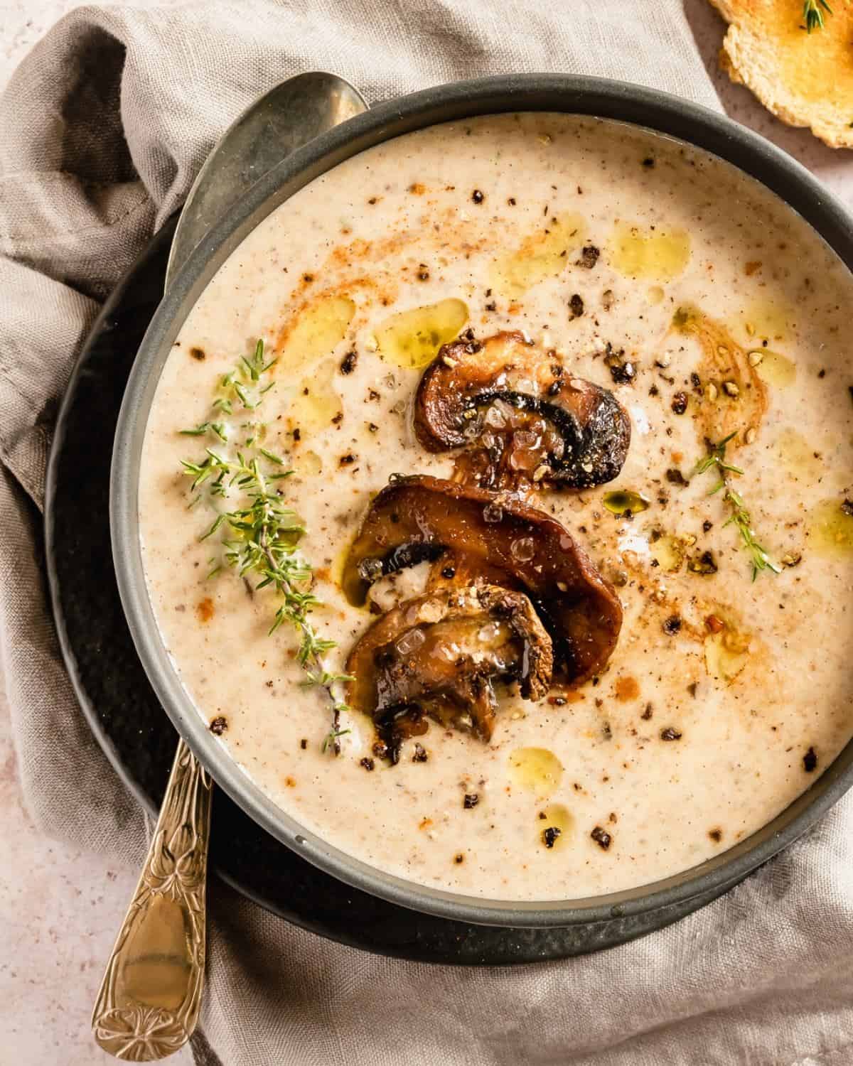 A view from above Mushrooms soup in a dark bowl on a beige linen and marble table. The dish is garnished with Mushroom and black pepper. On the side a spoon