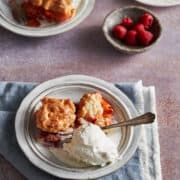 Slice of Peaches and Raspberries Cobbler in a white plate. The slice shown all fruit baked inside. It is served with some ice-cream