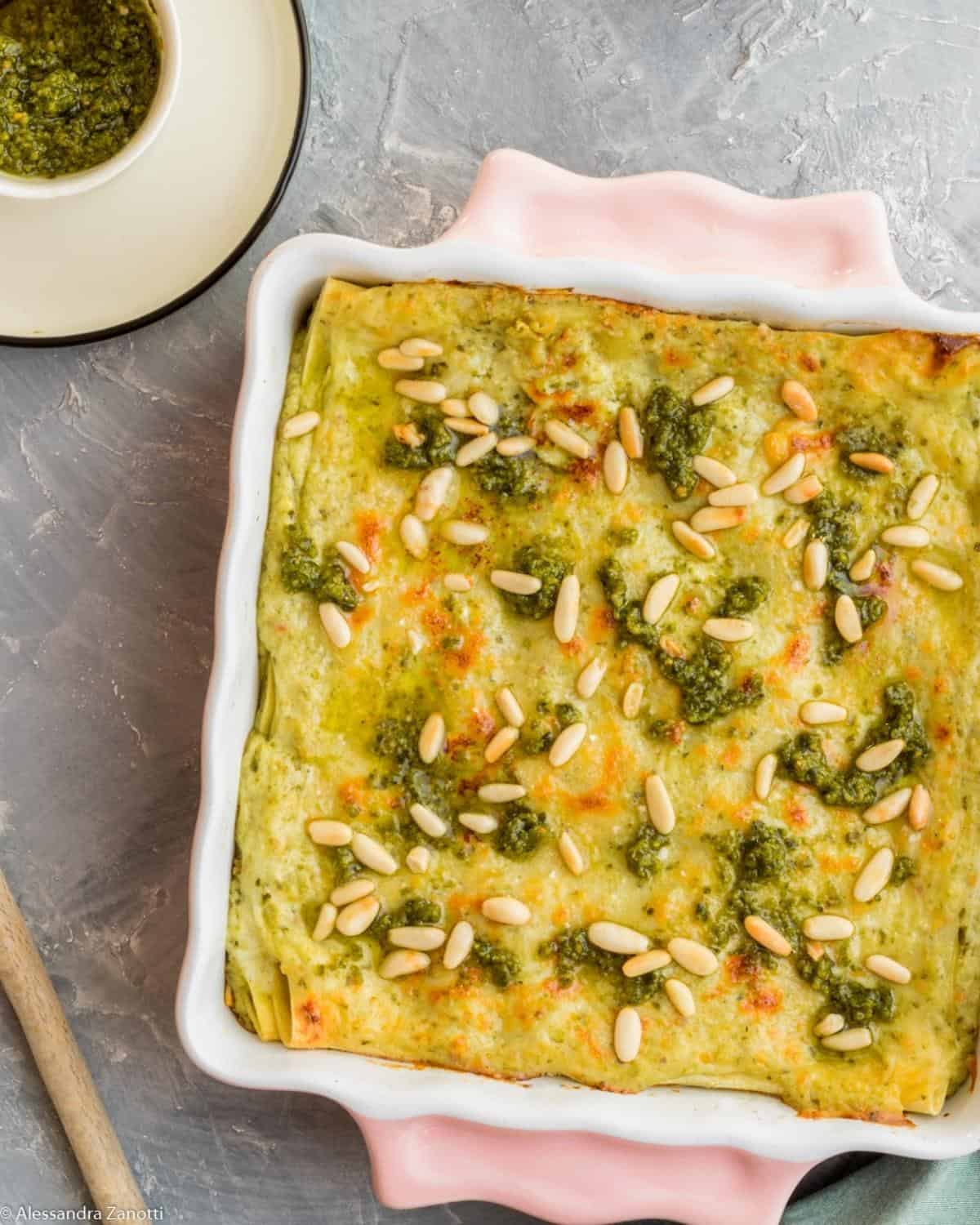 A close up from above of Pesto Lasagna in a pink pan. It is topped with pesto sauce and pine nuts