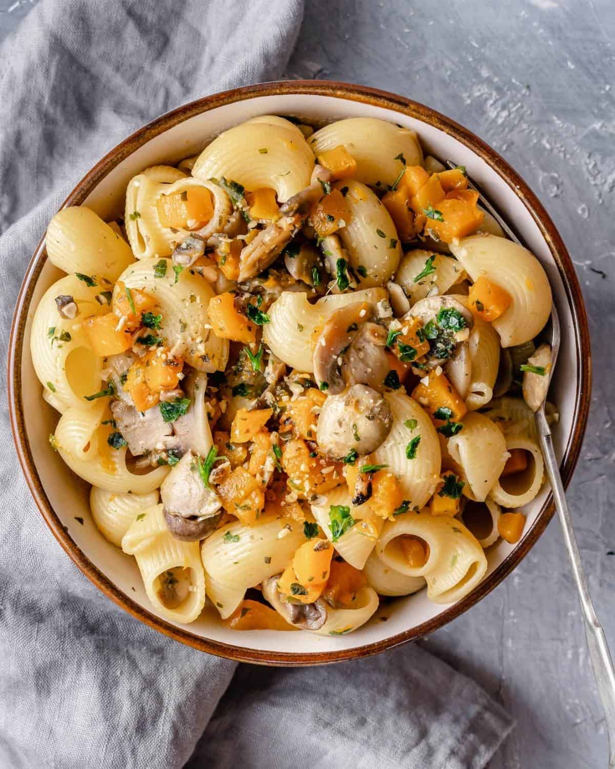 A close up of a pasta bowl with a fork showing pasta with pumpkin cubes, mushrooms, seeds and fresh herbs
